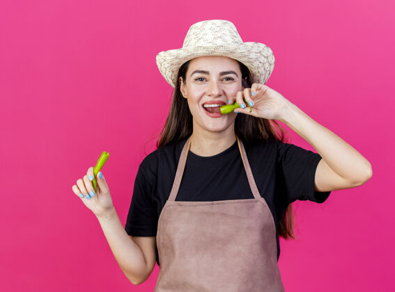 园艺微笑美丽的园丁女孩穿着制服 戴着园艺帽 在粉红色的背景上尝试辣椒隔离花园微笑胡椒