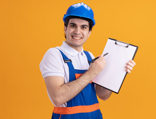 年轻人年轻的建筑工人穿着建筑制服 戴着安全帽 手里拿着写字板 手里拿着钢笔 面带微笑地站在橙色的墙上 看着前方微笑安全脸