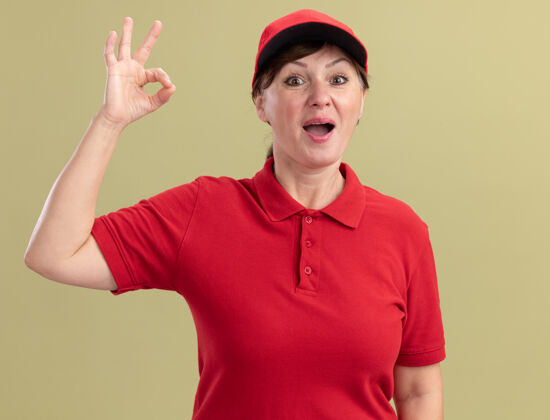 欢呼身穿红色制服 头戴帽子的中年女送货员站在绿色的墙上 面带微笑 脸上挂着“ok”的牌子表演年龄女人
