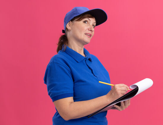 思考身穿蓝色制服 戴着帽子 拿着剪贴板和铅笔的中年女送货员站在粉红色的墙上沉思着铅笔站着制服