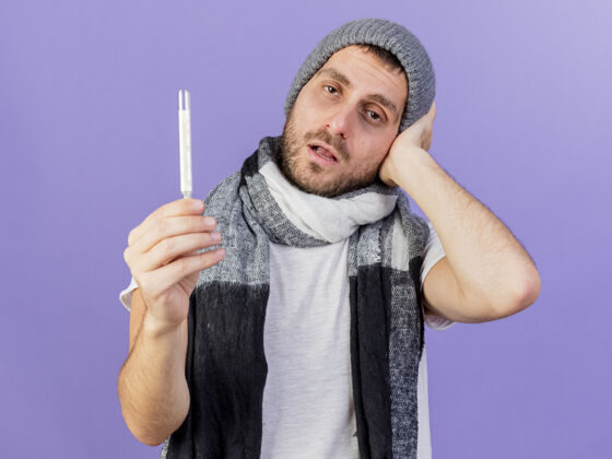 体温计戴着带围巾的冬帽 拿着体温计 手放在紫色背景上孤立的头上的未出狱的年轻病人头年轻人围巾