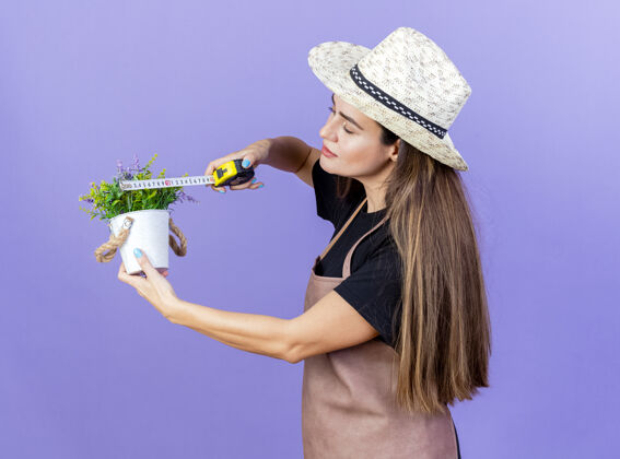 女孩穿着制服的漂亮园丁女孩戴着园艺帽 用卷尺在蓝色背景上测量花盆里的花花帽子花盆