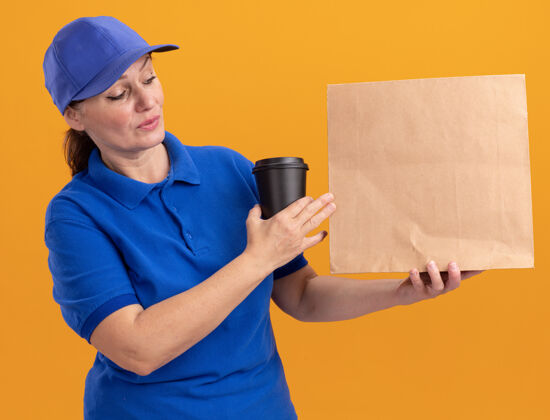 包装身着蓝色制服 戴着帽子 拿着纸包和咖啡杯的中年女送货员站在橙色的墙上 表情严肃咖啡交货年龄