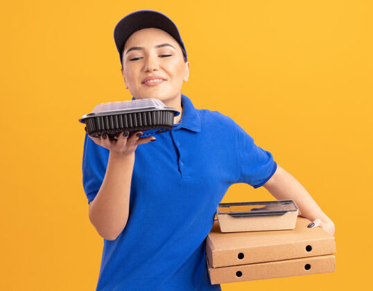 食物身着蓝色制服 头戴鸭舌帽 手持披萨盒和食品包装的年轻女送货员站在橙色的墙上 散发着怡人的香气制服立场吸入器