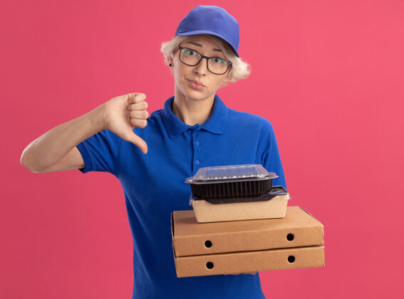 展示身穿蓝色制服 戴着眼镜 拿着比萨饼盒和食品包的年轻送货员 表情悲伤 在粉红色的墙上竖起大拇指盒子表情眼镜