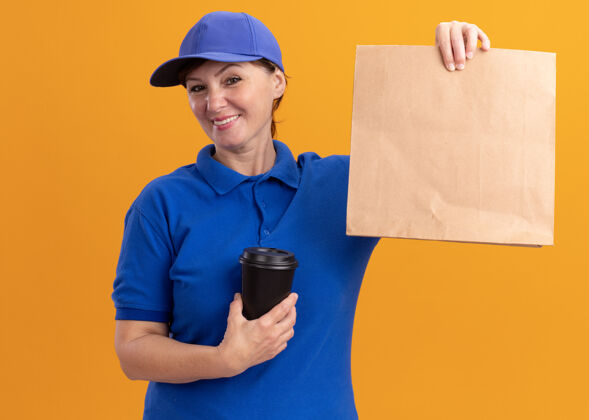 帽子快乐的中年送货员 身着蓝色制服 戴着帽子 展示着纸包 手里拿着咖啡杯 微笑着站在橙色的墙上看着前面微笑送货咖啡