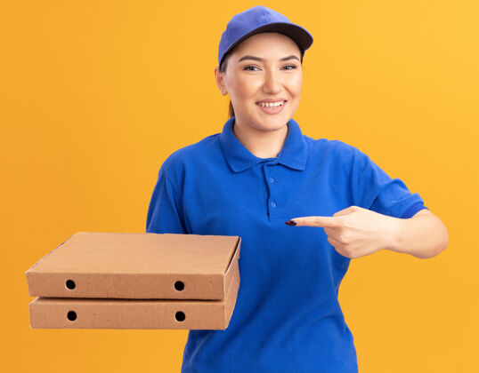 站着身着蓝色制服 戴着帽子 拿着披萨盒 用食指指着披萨盒 快乐地站在橙色的墙上 微笑着看着前面目录送货制服