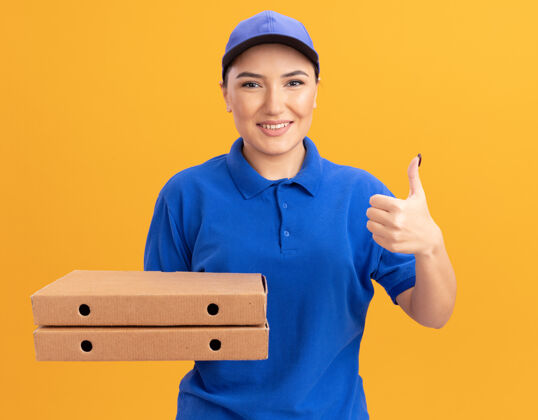 拇指身穿蓝色制服 头戴鸭舌帽的年轻送货员站在橙色的墙上 微笑自信地竖起大拇指 看着前面的比萨饼盒展示微笑自信