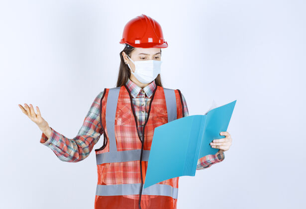 女人女建筑工程师戴着安全面具 戴着红色头盔 手里拿着一个蓝色的文件夹 看上去很困惑 很体贴报告姿势年轻人
