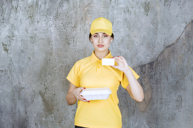 年轻人穿着黄色制服的女快递员送了一个白色的外卖盒 并出示了她的名片女人运输食物