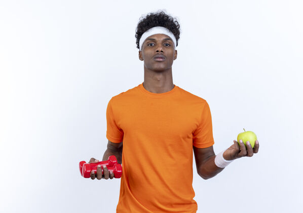 相机看着镜头年轻的美国黑人运动男子戴着头带和手环举着哑铃与苹果隔离在白色的背景头带运动穿