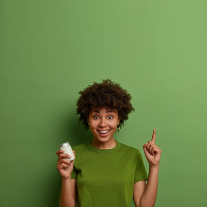 肖像快乐的黑皮肤女孩引领健康的生活方式 保持健康 早餐拿着一罐有机酸奶 用食指向上指 展示适合你的营养的食物或产品饮食容器早餐