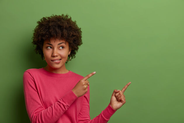 工人好奇的非裔美国妇女咬嘴唇的室内镜头 看到一些好东西 在绿色墙上的空白处指指点点 显示您的产品 公司横幅或促销的地方手势咬表演