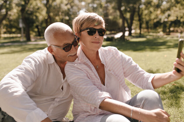 白色戴太阳镜的金发女郎 条纹时髦的衬衫和牛仔裤坐在草地上 和穿着白衬衫的胡子男人在户外自拍关系成人两个