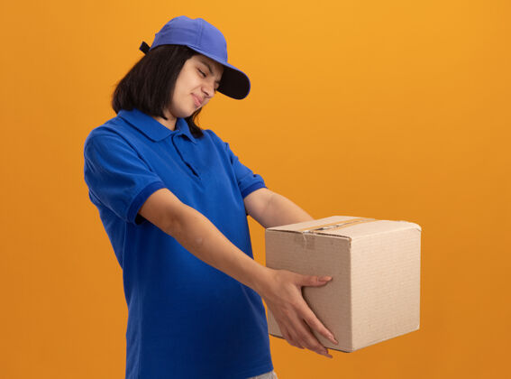 恶心身穿蓝色制服 戴着帽子 拿着纸板箱的年轻送货女孩站在橙色的墙上 带着厌恶的表情看着它女孩年轻制服
