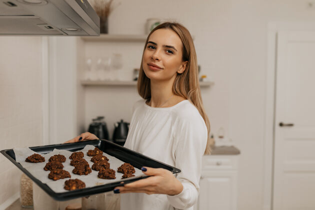 积极可爱可爱的女人 浅棕色头发 穿着白衬衫 手里拿着装饰饼干在厨房里平衡爱好快乐