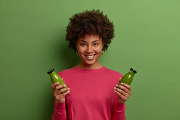 室内快乐微笑的黑皮女人肖像拥有健康自然的生活方式 拿着两瓶绿色蔬菜冰沙 有适当的营养 享受减肥饮料 穿着玫瑰色的套头衫 微笑新鲜生物卷曲