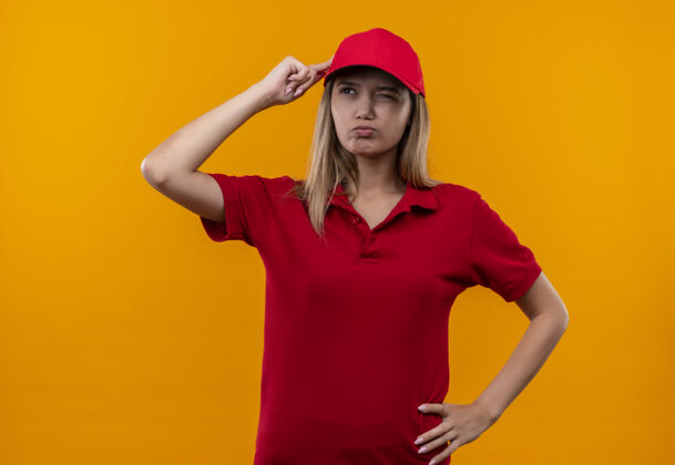 交货看着身边迷茫的年轻送货女孩穿着红色制服 戴着帽子 手指贴着头孤立在橙色的背景上穿帽子制服
