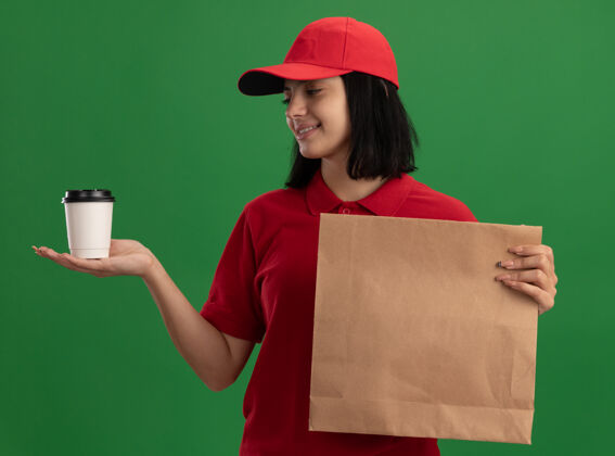 包装身穿红色制服 头戴鸭舌帽的年轻送货小姐手拿纸包和杯子 面带微笑地看着杯子站在绿色的墙上年轻站着递送