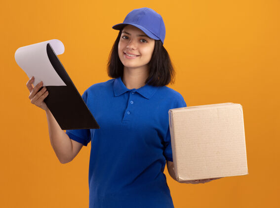 脸身穿蓝色制服 头戴帽子的年轻送货女孩站在橙色的墙上 手里拿着纸板箱和剪贴板 脸上挂着微笑的空白页页面制服空白