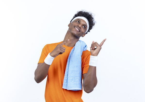 肩膀看着面带微笑的年轻美国黑人运动男子戴着头带和腕带点在一边与毛巾在肩上隔离在白色背景与复制空间年轻点毛巾