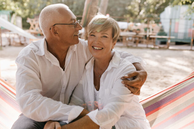 女人一个快乐的女人 一头金发 穿着白色衣服 坐在吊床上 和一个戴着眼镜微笑的男人拥抱在沙滩上退休两个年龄