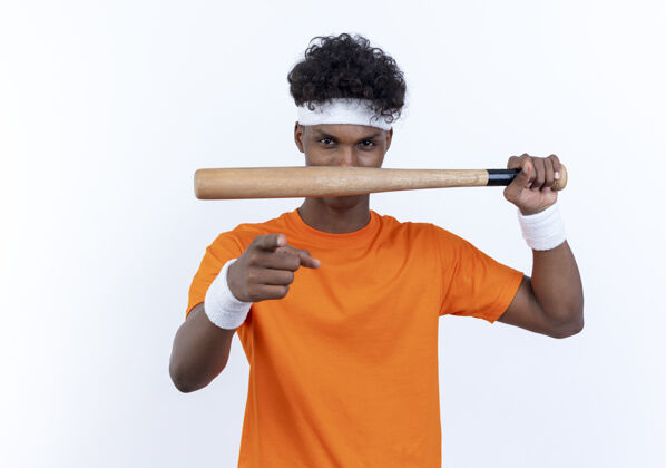 运动看着镜头 年轻的美国黑人运动男子戴着头带和腕带拿着球棒 在白色背景上向你展示孤立的手势腕带表演头带