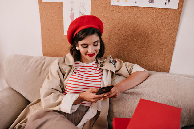 成人戴着鲜艳贝雷帽的快乐女士在智能手机里聊天穿着时髦衣服的漂亮女人正坐在沙发上微笑着礼品盒黑发红唇