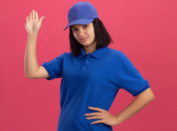 手身穿蓝色制服 头戴鸭舌帽的年轻送货小姐自信地站在粉色的墙上挥手致意立场年轻信心