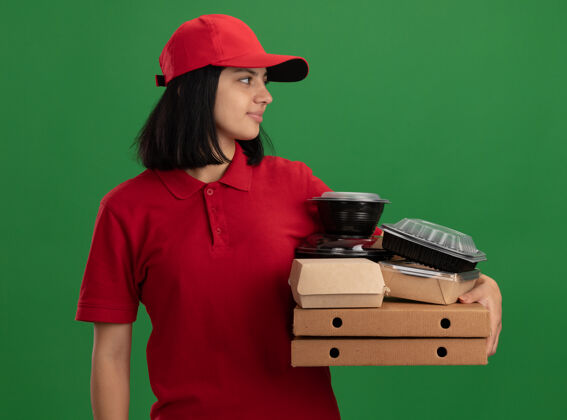 脸身穿红色制服 头戴鸭舌帽 手持披萨盒和食品包的年轻送货员站在绿色的墙上 面带微笑地看着一旁食物微笑送货