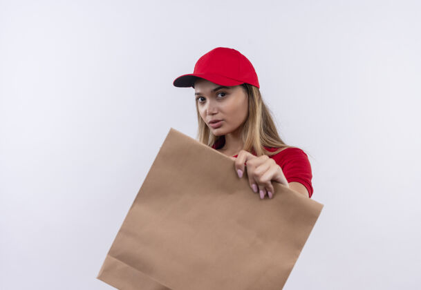 包看着镜头 年轻的送货女孩穿着红色制服 戴着帽子 手里拿着纸袋 隔离在白色背景上帽子年轻人拿着
