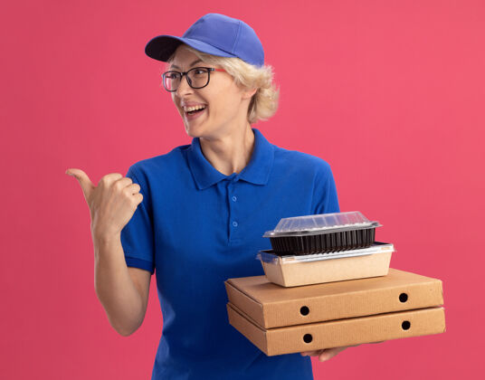 壁板身穿蓝色制服 戴着眼镜 手持比萨饼盒和食品包的年轻送货员微笑着 用食指指着粉色墙壁的一侧微笑眼镜手指