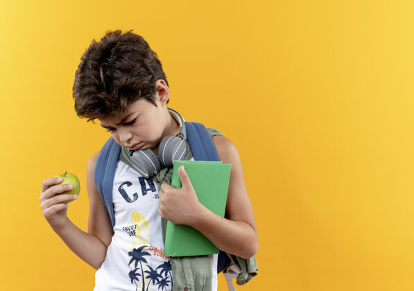 耳机看着那个戴着背包和耳机 拿着苹果和书 被隔离在黄色背景上 留着复印空间的悲伤的小男孩苹果背抱