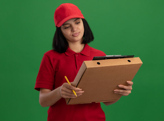 脸身穿红色制服 头戴鸭舌帽的年轻送货女孩拿着比萨饼盒和铅笔的剪贴板看着剪贴板 脸上带着微笑站在绿色的墙上站着女孩帽子