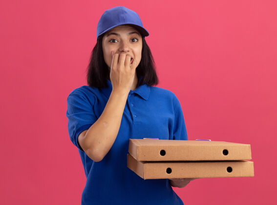 披萨穿着蓝色制服 戴着帽子 手里拿着披萨盒的年轻送货女孩站在粉红色的墙上紧张地咬着指甲盒子咬女孩