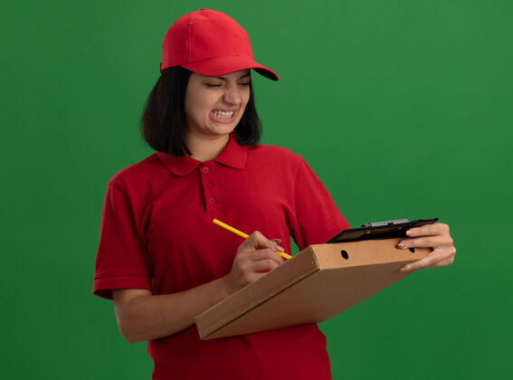 剪贴板身穿红色制服 头戴鸭舌帽 拿着比萨饼盒和铅笔的剪贴板的年轻送货员站在绿色的墙上写着困惑和不快的东西铅笔拿着帽子