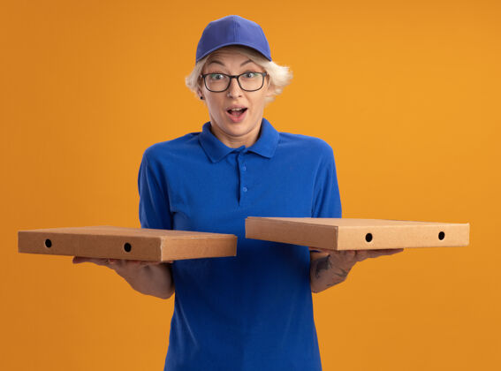 披萨身着蓝色制服 戴着眼镜 拿着比萨饼盒的年轻送货员在橙色的墙上惊讶不已惊喜帽子送货