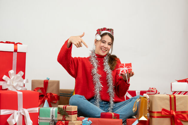 快乐前视图可爱的派对女孩 戴着圣诞帽指着她的礼物坐在礼物旁边周围礼物圣诞老人