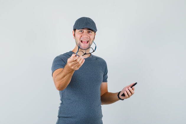 电脑摄像头一个戴着灰色t恤帽的年轻男人 手里拿着手机和眼镜 看上去很快乐电话隔离商务摄像头