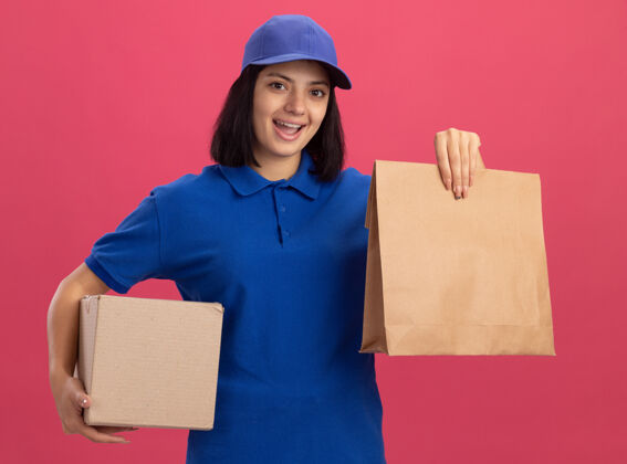 微笑身着蓝色制服 头戴鸭舌帽 拿着纸包和纸板箱的快乐的年轻送货女孩站在粉红色的墙上 开心地微笑着抱着送货站着