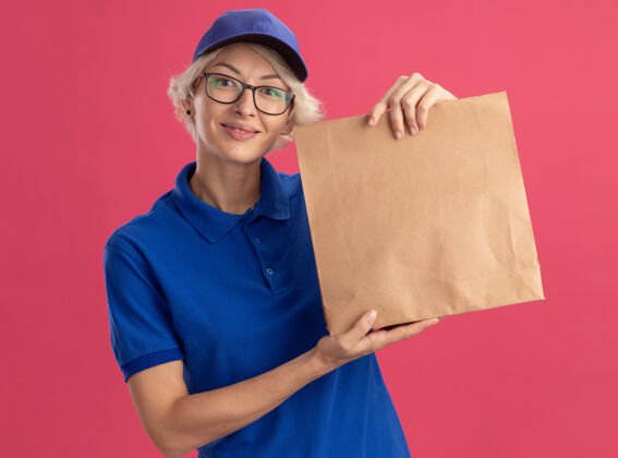 欢呼身穿蓝色制服 戴着眼镜 拿着纸包的年轻送货员在粉红色的墙上欢快地微笑着送货包装戴着