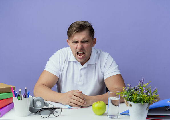 年轻愤怒的年轻英俊的男学生坐在书桌旁 蓝色背景上孤立着学习工具生气男课桌