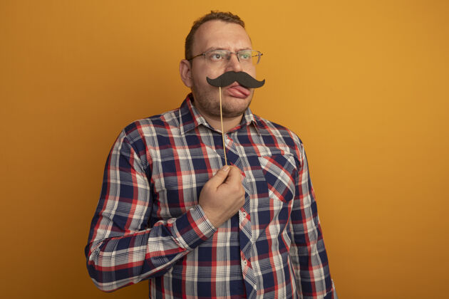搞笑一个戴着眼镜 穿着格子衬衫的男人站在橘色的墙上 手里拿着一把滑稽的小胡子 伸出舌头眼镜棍子舌头