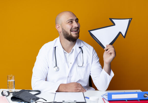 医疗年轻的秃顶男医生穿着医用长袍和听诊器坐在办公桌旁 手持医疗工具 手持橙色背景上孤立的方向标记工作方向长袍