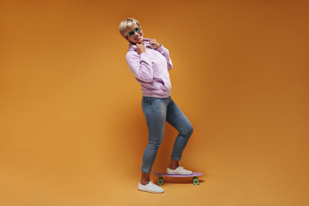 牛仔裤时髦的老妇人 金发碧眼 穿着粉色运动衫 牛仔裤和白色运动鞋 微笑着摆出滑板的姿势女衬衫中
