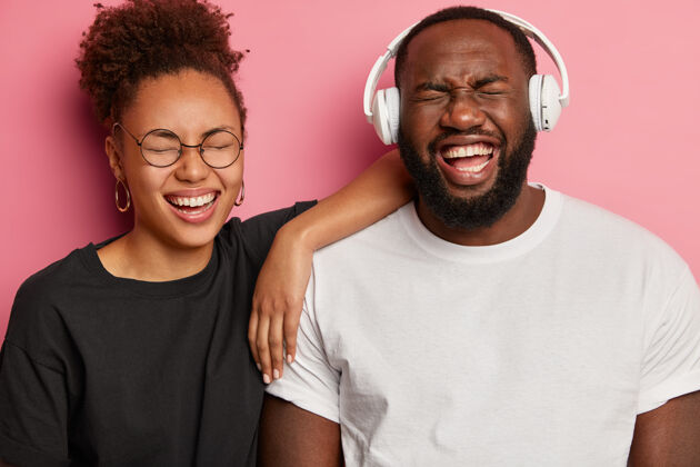 音乐喜出望外的美国黑人夫妇玩得很开心 戴着耳机听最喜欢的音乐 对一些积极的事情傻笑 穿黑白衣服 享受在一起的时光 隔离在粉红色的墙上年轻人欣喜若狂快乐