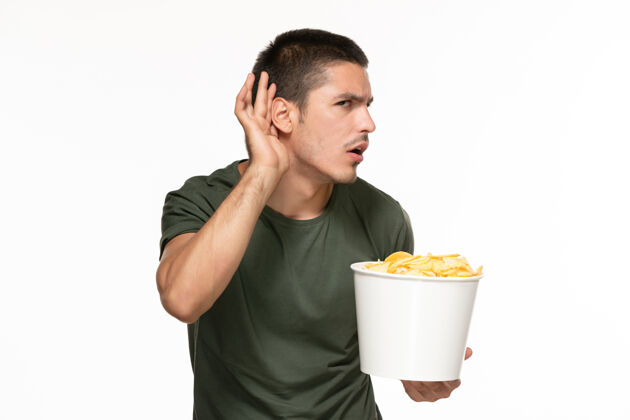 年轻男性正面图身穿绿色t恤的年轻男子拿着篮子和土豆 试图在白墙上听到孤独的享受电影享受成人电影