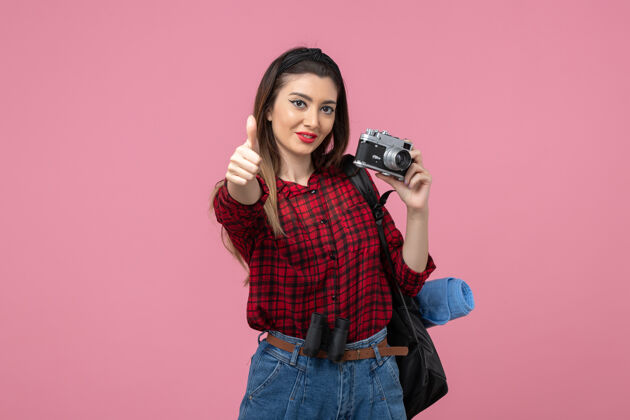牛仔裤正面图：穿着红色衬衫的年轻女性 在粉色背景上用相机拍摄模特照片裤子相机衣服