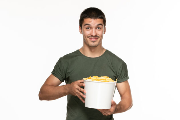 享受正面图身着绿色t恤的年轻男性 手拿土豆cips篮子 微笑着站在白墙上孤独地享受电影电影院成人景观杯子