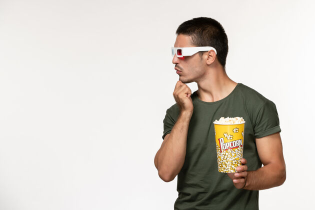 视图正面图身着绿色t恤的年轻男性手持爆米花in-d太阳镜在白色墙上看电影孤独电影院男性电影青少年电影电影